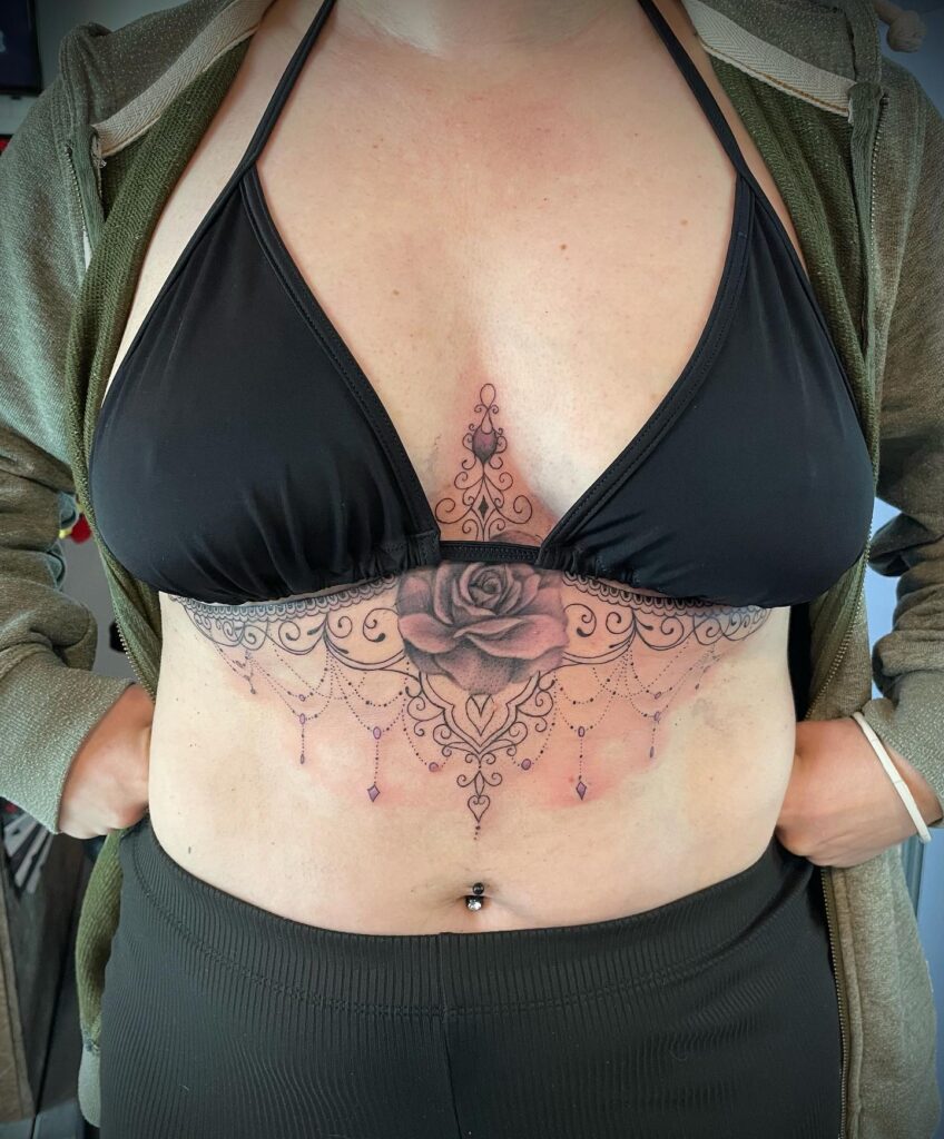 Under-breast tattoo featuring a detailed rose with ornamental designs and hanging chandelier accents