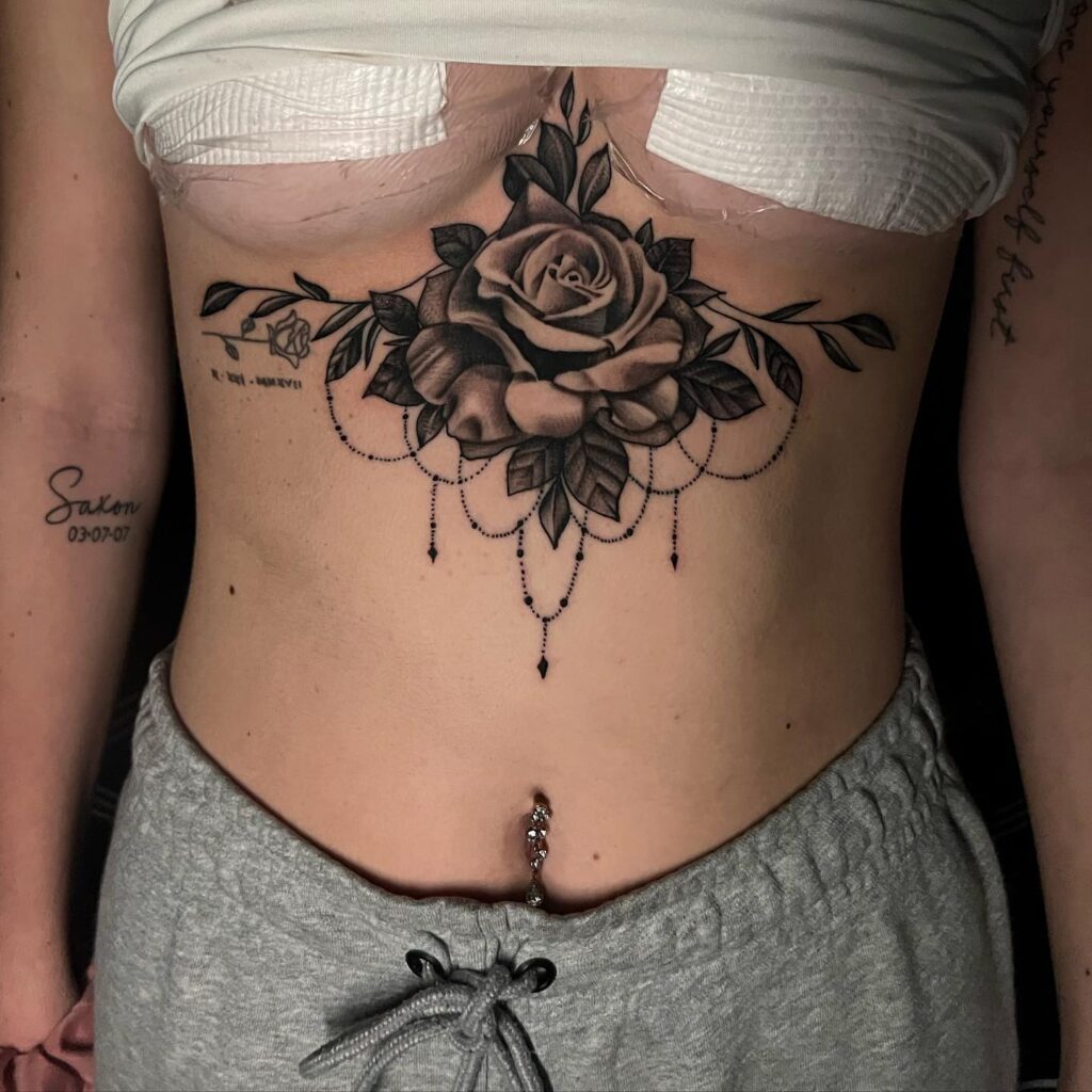 Under-breast tattoo featuring a large, realistic rose with detailed shading and hanging beaded accents, creating a feminine and elegant design