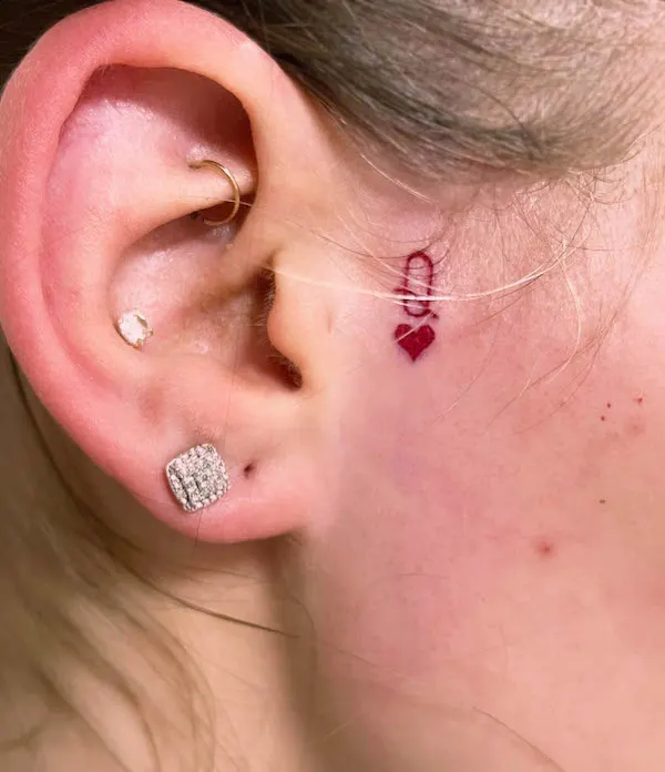 Small red heart tattoo behind the ear with a delicate symbol above, symbolizing love, emotions, and personal connection