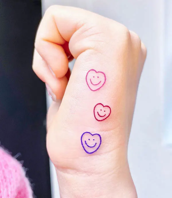 Three colorful smiling heart tattoos in pink, red, and blue ink on side of hand