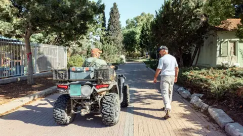 Maya Meshel / BBC Rami and Simon, both with their backs to the camera. Rami is on an all-terrain vehicle. Simon is walking to the right of him.