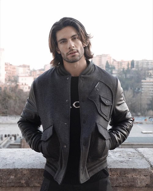 A man with medium-length hair wearing a dark varsity jacket, standing against a cityscape background, exuding a casual and stylish look.