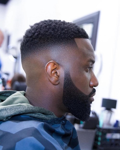 Man with a fade crew cut hairstyle and a well-groomed beard, wearing a camouflage jacket in a barbershop