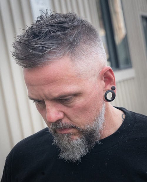 Man with a textured crew cut hairstyle, featuring a fade and gray beard, wearing a black t-shirt and ear gauges