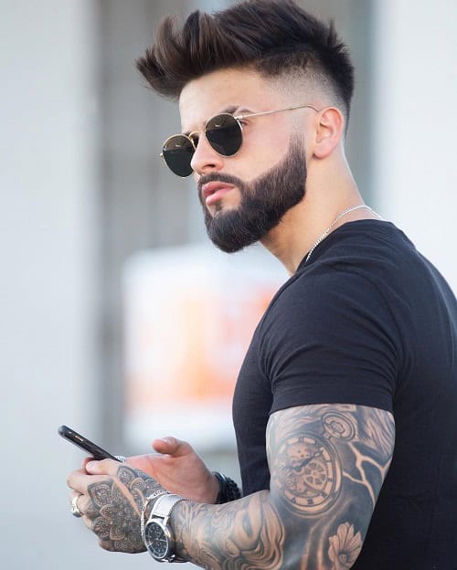 A man with a high-fade hairstyle, a well-groomed full beard, wearing sunglasses and showcasing sleeve tattoos, while holding a phone