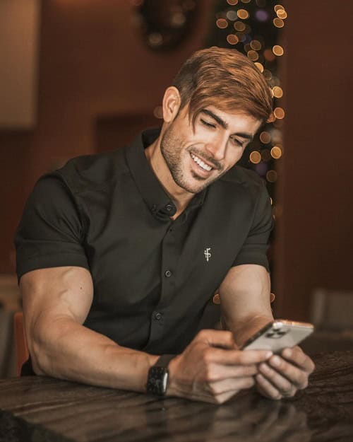 A man with a stylish blonde side-part haircut, wearing a black button-up shirt, smiling while holding a smartphone, seated indoors with a festive background