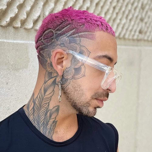 A man with a vibrant pink buzzcut hairstyle and intricate side head tattoos, wearing clear glasses and a nose ring, standing against a textured wall.