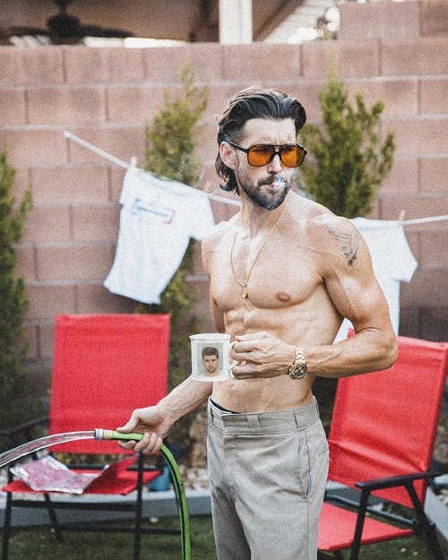 Shirtless man with a slicked-back mullet haircut, wearing sunglasses and holding a coffee mug while watering the garden