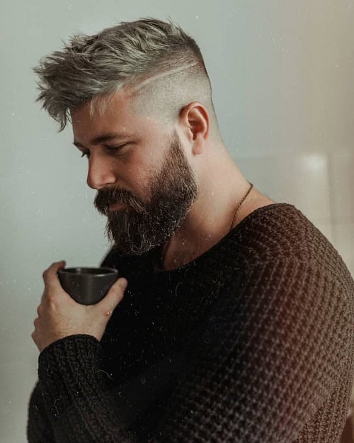 Man with a textured crew cut hairstyle and a thick beard, featuring a shaved line design, wearing a black sweater