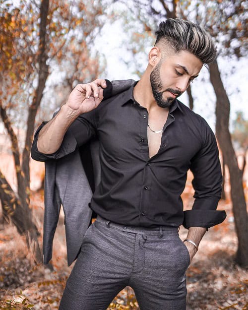 A man with a stylish modern undercut hairstyle, wearing a black shirt and gray pants, holding a jacket over his shoulder while standing in an outdoor setting with trees in the background.