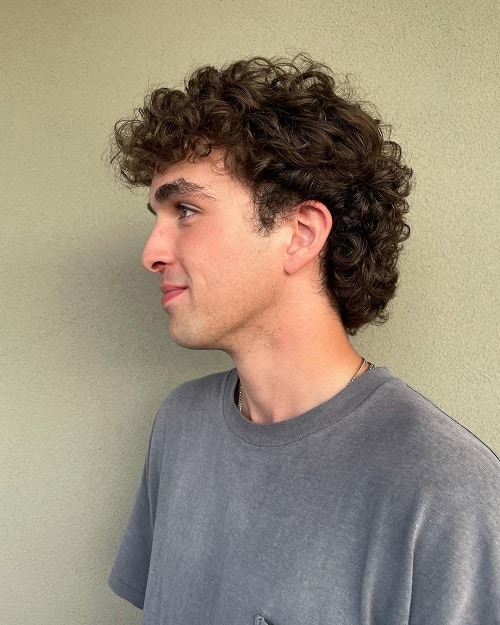 Side view of a man with a short curly mullet haircut, featuring soft curls on top and a subtle mullet at the back