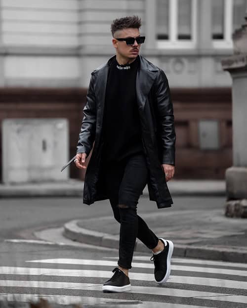 A man with a spiky modern haircut, wearing sunglasses, a black leather jacket, and black jeans, walking confidently across a city street in an all-black urban outfit