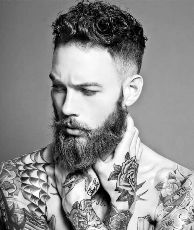 Portrait of a man with a curly drop fade haircut, full beard, and detailed tattoos on his chest and arms.