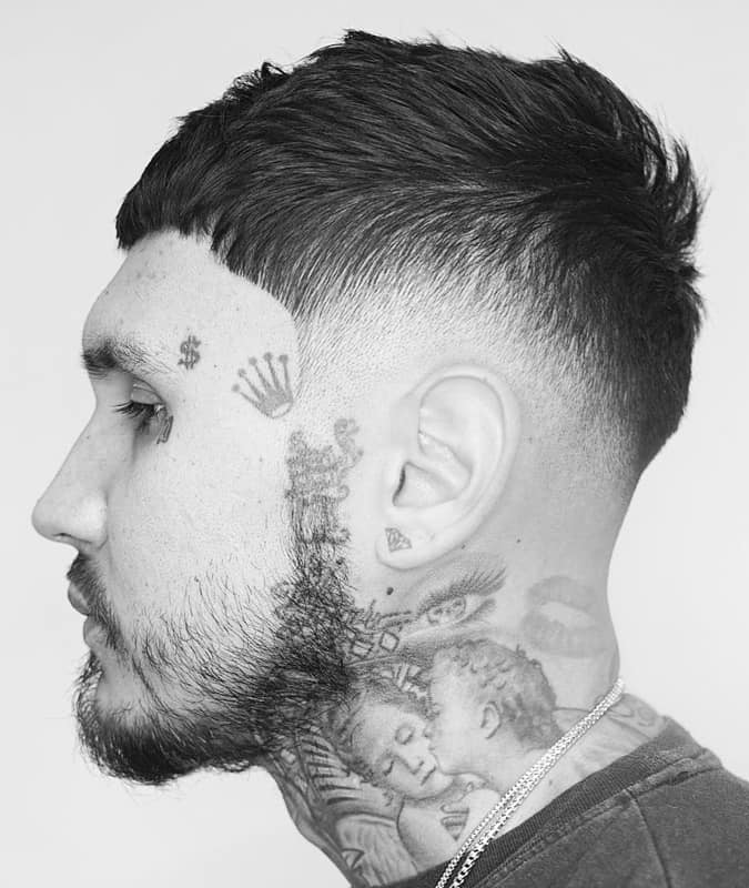 Side profile of a man with a textured drop fade haircut, short fringe, and prominent facial and neck tattoos.