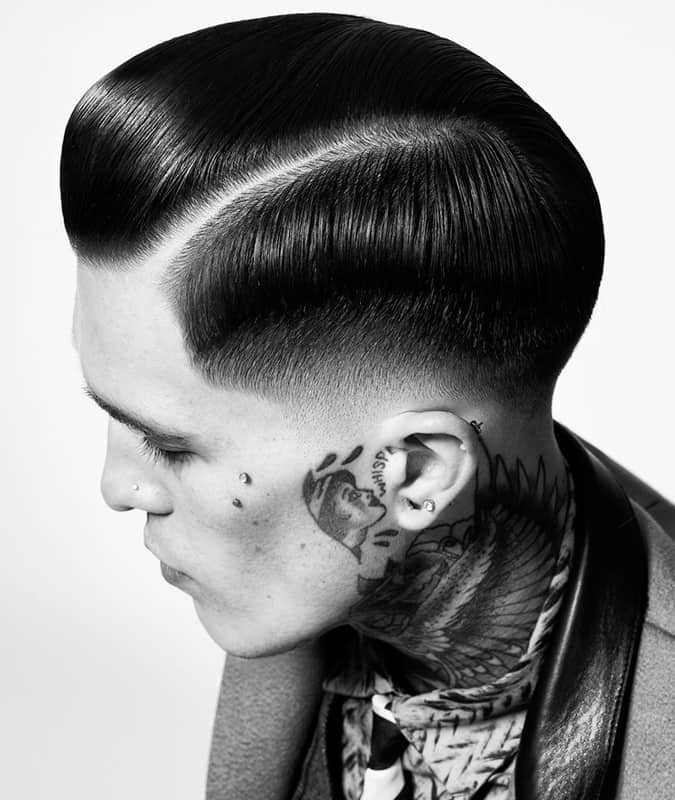 Top view of a man with a slicked-back drop fade haircut featuring a hard part and prominent face and neck tattoos.