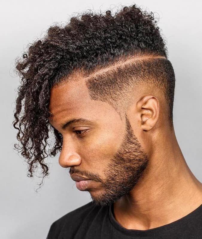 Side profile of a man with a curly drop fade haircut, hard part, and a neatly shaped beard.