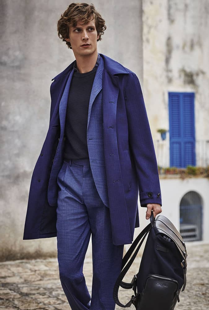 Man in a monochromatic blue ensemble featuring a checkered suit, matching trench coat, and a black bag, showcasing a sophisticated travel-ready look