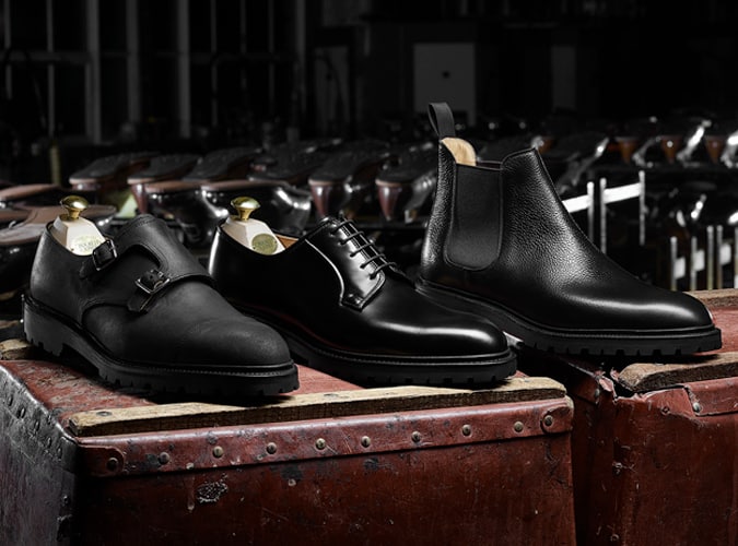 Three types of black men's leather shoes displayed on vintage trunks: double-monk strap, Derby, and Chelsea boots, showcasing classic styles with rugged soles