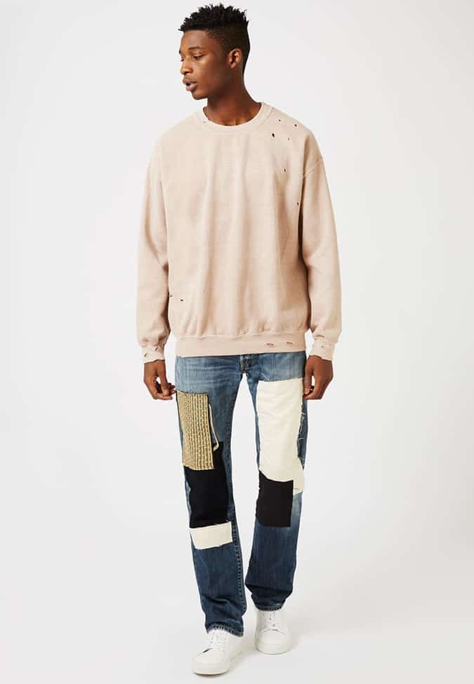 Man in a beige distressed sweater and patchwork jeans, styled with white sneakers for a minimalist grunge-inspired look