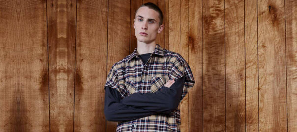 A young man wearing a modern grunge-inspired outfit with a plaid flannel shirt over a black long-sleeve shirt, standing against a wooden wall background