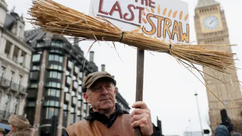 Reuters A man taking part in the protest wears a leather waistcoat and flat cap. He holds a sign which reads 'the last straw' and which has straw attached to it. In the background is Big Ben