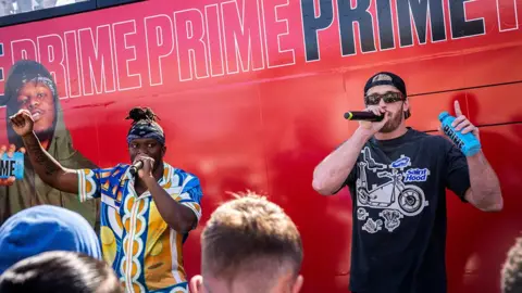 Reuters KSI and Logan Paul stand in front of a red bus marked repeatedly with the word "prime", apparently in front of a crowd (only a few heads visible). Both are talking through mics and Logan Paul is holding a blue bottle of Prime. KSI is wearing a blue and yellow Hawaiian shirt and a bandana; Paul is wearing a baseball cap back to front, and a black t-shirt with the insignia "Saint Hood"