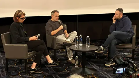 Bianca Cline and Joel Souza sit in armchairs in front of the screen and speak into microphones. A third person listens to them. There is a table in front of them with bottles of water.