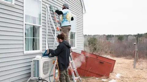 Getty Images Workmen fit a heat pump to the to the clapboard exterior of a house.