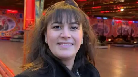 Nihal Nihal with dark hair and a fringe, smiling at the camera, standing in front of bumper cars at a fairground