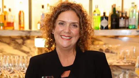 Getty Images Hannah Ingram-Moore appears in front of a bar, with glasses and bottles behind her. She wears a black jacket. Her hair is curly and she is wearing red lipstick.