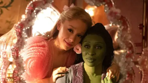 Universal Ariana Grande and Cynthia Erivo stare into a pink mirror dressed as their Wicked characters Glinda and Elphaba.