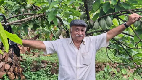 George Matthew George Matthew wearing a flat cap stands in front of a cocoa tree, with cocoa pods hanging from branches.