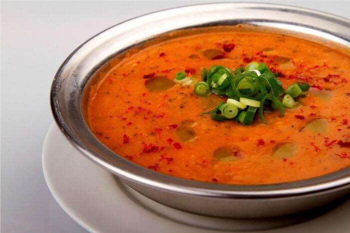 A bowl of Turkish lentil soup garnished with green onions and chili flakes, served in a metal bowl.