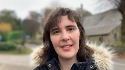 BBC The picture shows Dee Goddard. She is wearing a winter coat and smiling. She is pictured in the Derbyshire Dales village of Curbar