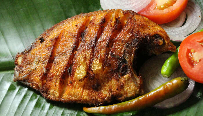 Kerala-style Karimeen Pollichathu, grilled pearl spot fish served on a banana leaf with sliced tomatoes, onions, and green chilies.