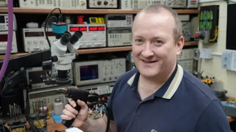 Mark Maher Mark Maher holding a soldering device, behind him are shelves full of electrical equipment