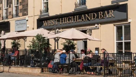 Getty Images Front entrance of West Highland Bar in Portree, Isle of Skye. The venue has a black and gold sign with a number of tables, umbrellas and patrons outside on a sunny day. 