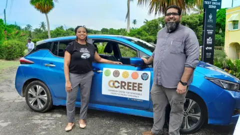 Dr Legena Henry stands next to a blue car with a sign reading "Caribbean Centre for Renewable Enert and Enery Efficiency" on its door. 