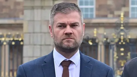 Colin McGrath with greying hair and a beard stares to his right with a stern look on his face. He is wearing a blue/grey suit, white tie and brown tie.