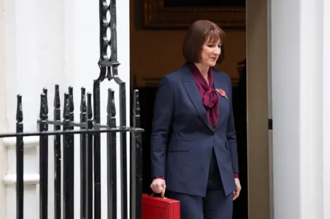 PA Media Rachel Reeves coming through the door of 11 Downing Street holding her red Budget box and wearing a blue suit, a burgundy top and a red poppy. There are black railings in front of the white building.