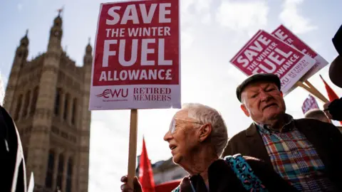 EPA Two elderly protesters hold red placards saying "Save the winter fuel allowance" outside the House of Parliament, which can be seen in the background. One is looking to the left and the other is staring at the camera, wearing a flat cap and a checked shirt.