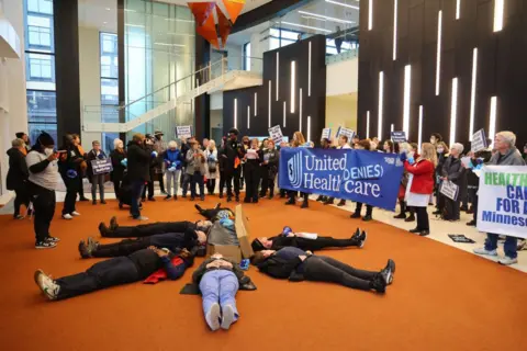 Getty Images Protesters with People's Action lay in circle at health insurance giant UnitedHealth Group's headquarters holding signs saying "UnitedHealth denies care"