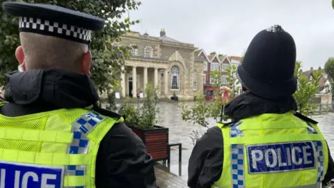 The back of police officers heads standing in front of the Guildhall in Salisbury. Its a very big grand building, with pillars of stone with a large square out the front.