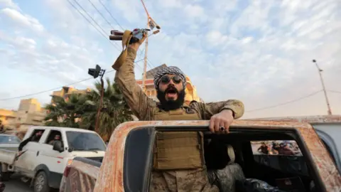 A man in all brown military clothing leans out of the door of a rusty red car, holding a weapon aloft.