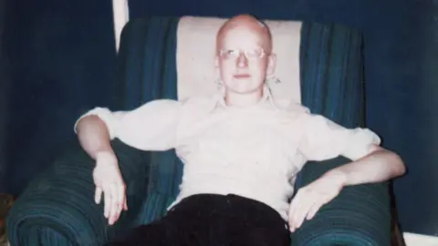 Family photo A teenage Dave Myers sits on a blue armchair. He has no hair and is wearing a white shirt, dark trousers and glasses. He looks serious and is staring at the camera.