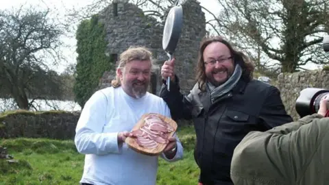 Family photo Si and Dave stand posing for the camera. Si is dressed in a white top and is smiling. He is holding a tray of bacon. Dave stands next to him and is wearing a leather jacket. He has a big grin on his face and is holding a frying pan above his head.