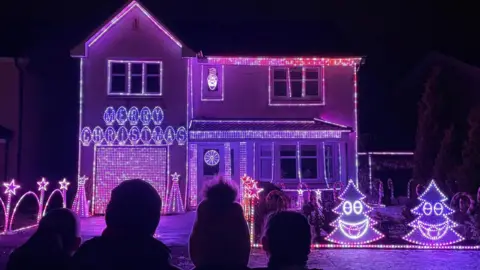 Gary Gray A two-storey home illuminated in bright pink and purple Christmas lights. There are small LED Christmas trees with smiley faces and the driveway is bordered by brightly lit archways and stars.