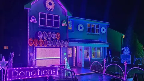 Gary Gray A two storey house brightly illuminated with LED Christmas lights in shades of blue and pink. There is a sign above the garage door that reads Merry Christmas. In the front corner of the garden is a pink sign which reads "Donations". 