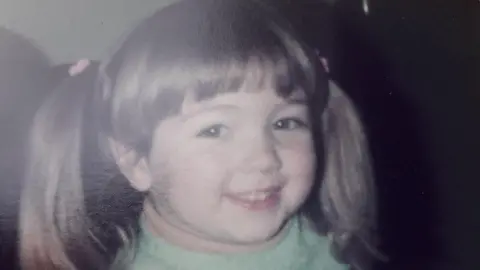 Tricia Hayes Steff when she was young with bunches in her brown hair. She is smiling at the camera and has a green jumper on.