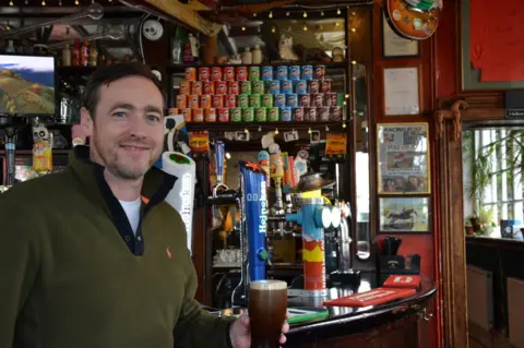 Citystack London Pub Collection A man in a khaki fleece is holding a pint of beer and smiling at the camera. He is stood in front of a bar that has bar taps, tubs of Pringles and posters on the wall